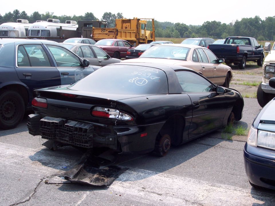 Black sports car with missing wheels and bumper damage, parked in a junkyard with other vehicles