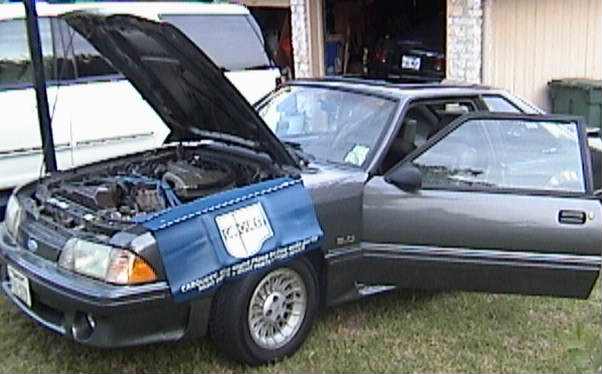 Gray Ford Mustang 5.0 hatchback with hood up in a yard, blue car cover hanging on the fender