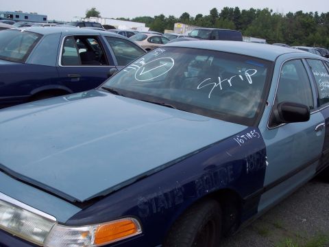 1991 Ford LTD Crown Victoria - Two police cruisers are parked in a lot with other vehicles. One has markings on the windshield and fender.