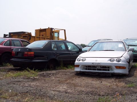 1991 Ford LTD Crown Victoria - Several vehicles and heavy equipment are parked in a junkyard under a cloudy sky.