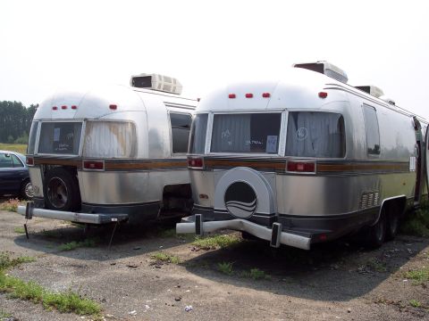 1991 Ford LTD Crown Victoria - Two Airstream trailers are parked side-by-side in an outdoor lot, with one showing damage to its rear bumper.