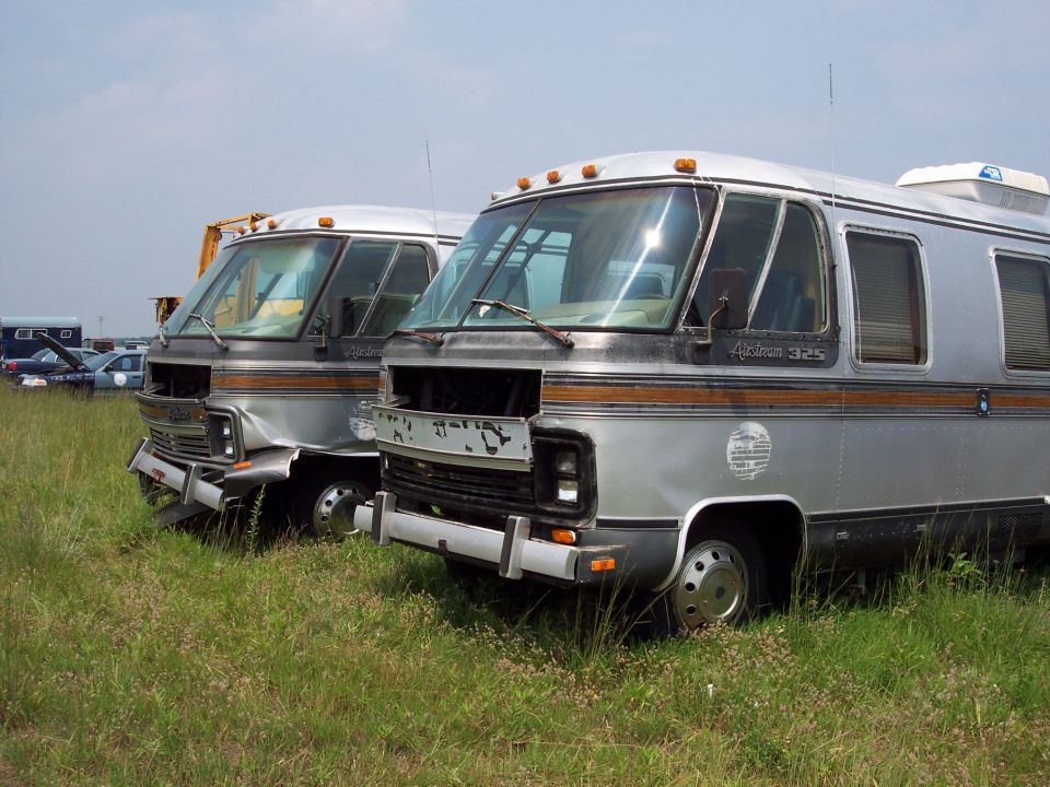 Two silver motorhomes parked in a grassy field, one with a damaged front bumper