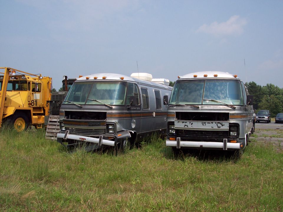 Two RVs parked in a grassy lot next to a yellow bulldozer. One RV has 'Olympian 305' on its side