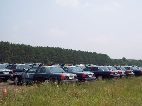 1991 Ford LTD Crown Victoria - A large group of police cruisers are parked in a grassy lot with a line of trees in the background.
