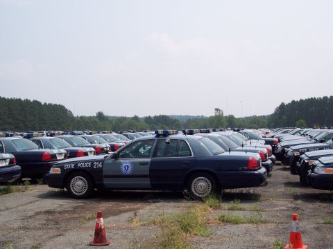 1991 Ford LTD Crown Victoria - A fleet of Ford Crown Victoria LTD police cars, including one marked 'STATE POLICE 214', are parked in a lot under a cloudy sky.