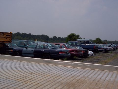 1991 Ford LTD Crown Victoria - A row of cars sits in a junkyard under a cloudy sky. The foreground shows a metal platform.