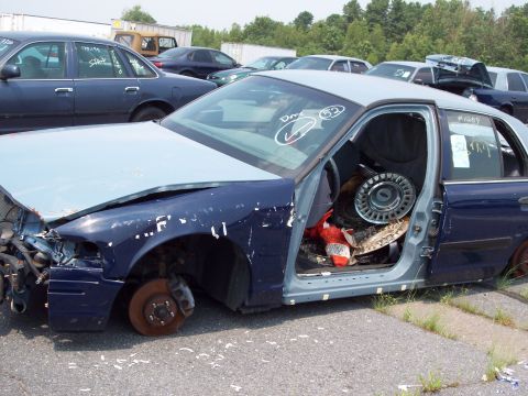 1991 Ford LTD Crown Victoria - A damaged Ford Crown Victoria sits in a junkyard with other vehicles visible in the background.