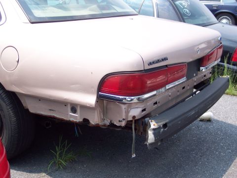 1991 Ford LTD Crown Victoria - Rear view of a damaged tan Ford Crown Victoria with its bumper detached, revealing rusted metal and wiring.