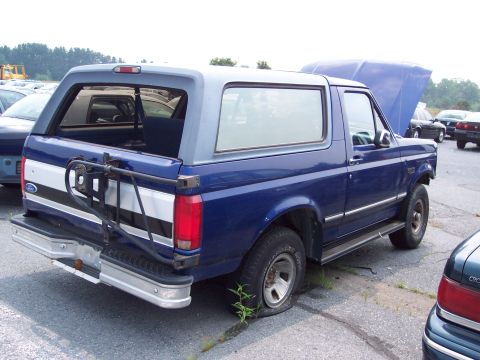1991 Ford LTD Crown Victoria - A blue Ford Bronco sits in a lot with its hood open and a flat tire. Another vehicle is partially visible on the right.