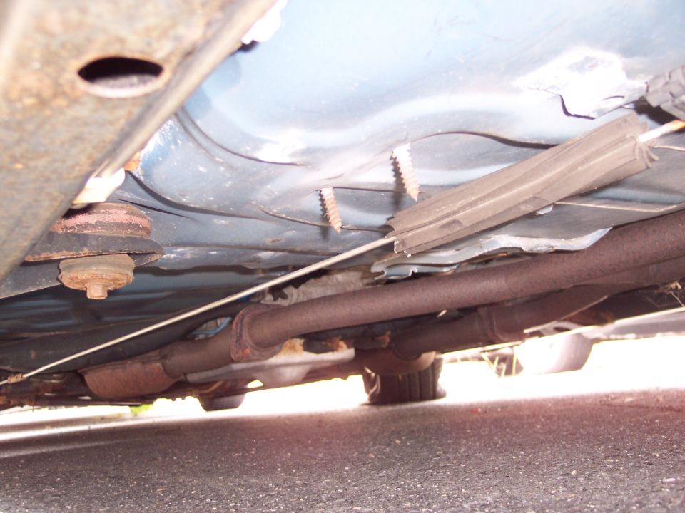 Underside view of a car, showing the exhaust system and frame. Two screws are visible in the body panel