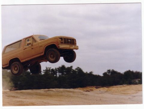 1987 Ford Bronco - A tan Ford Bronco is captured mid-air during a jump over a dusty terrain.