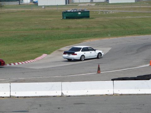 1992 Ford Taurus - A white sedan with a black trunk navigates a turn on a race track, with another vehicle partially visible on the left.