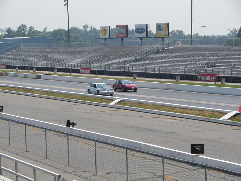 Two cars racing down a drag strip with empty bleachers in the background