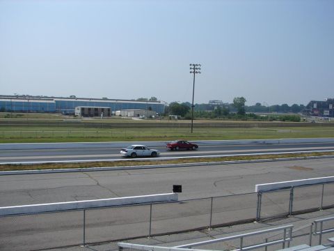 1992 Ford Taurus - Two sedans compete on a drag strip under a clear sky, with a large industrial building and stadium visible in the distance.
