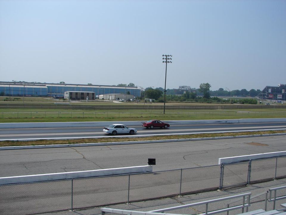 White and red sedans racing on a drag strip, with a large building and stadium in the background