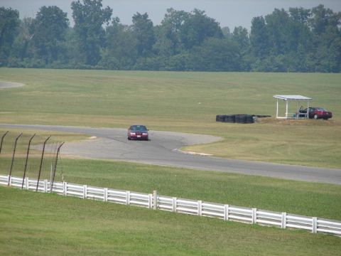1992 Ford Taurus - A red sedan navigates a turn on a race track, with another vehicle visible under a canopy in the distance.