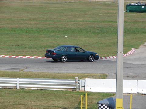 1992 Ford Taurus - A dark green sedan navigates a turn on a race track, with grassy infield and track barriers visible.