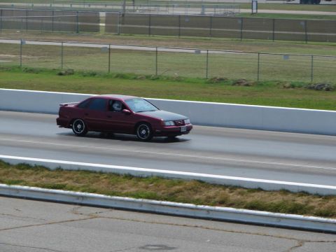 1992 Ford Taurus - A red 1992 Ford Taurus sedan with a black hood is seen driving on a race track.