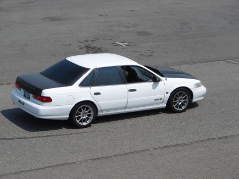 1992 Ford Taurus - A white 1992 Ford Taurus sedan with a black hood and custom wheels is viewed from a high angle on a paved surface.