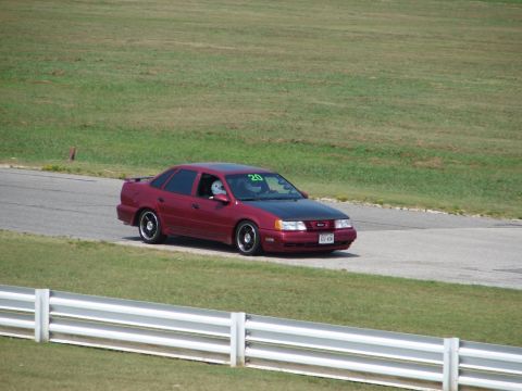 1992 Ford Taurus - A red sedan with a black hood is driving on a race track. The driver is wearing a helmet and the number 20 is visible on the windshield.