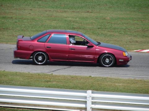 1992 Ford Taurus - A red 1992 Ford Taurus sedan with a black hood and racing wheels is seen on a race track.