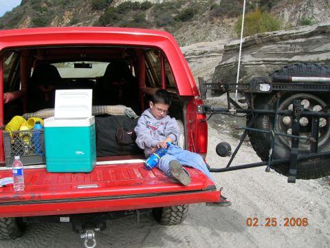 1985 Ford Bronco - A boy sits on the open tailgate of a red SUV, surrounded by camping gear and a cooler, with a spare tire mounted on the rear carrier.