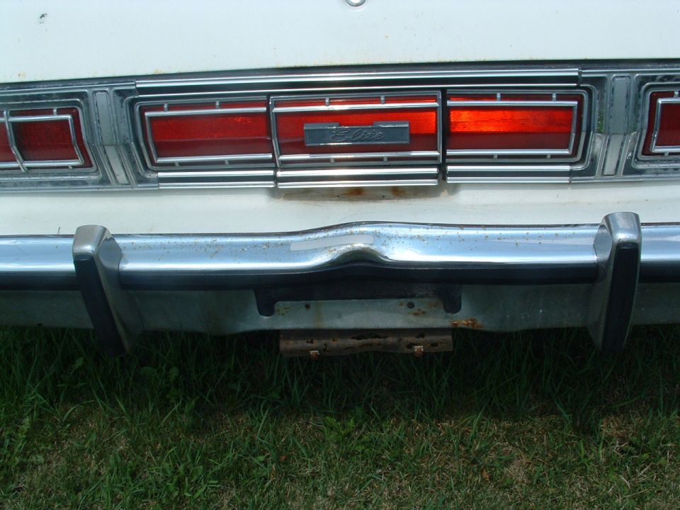 Rear of a white car showing a dented chrome bumper and a rectangular taillight with horizontal red lenses and a chrome trim piece