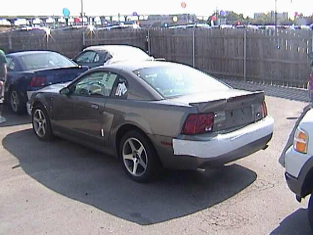 Gray Ford Mustang coupe rear three-quarter view on a car lot with wood fence