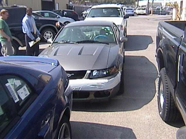 Front view of a silver Ford Mustang with hood scoops and tape on the bumper. A blue car is in the foreground