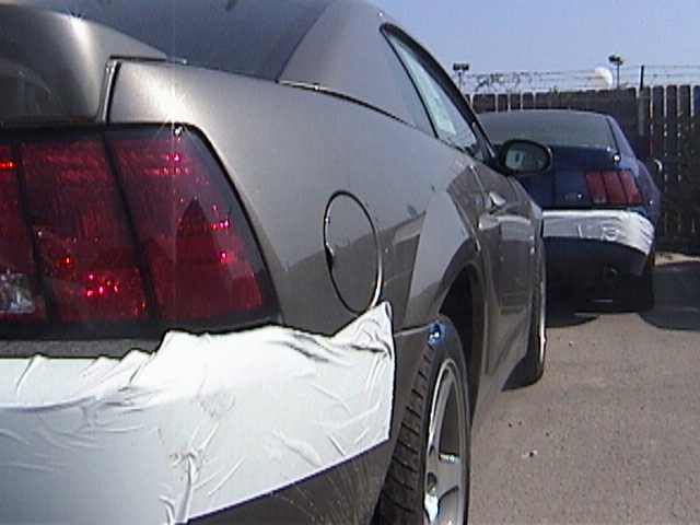 Gray Ford Mustang rear quarter panel with white protective wrap on bumper and fender. Another blue Mustang is parked behind it