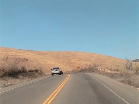 1971 Ford Bronco - A white pickup truck drives on a road with a dark car following, under a clear blue sky.