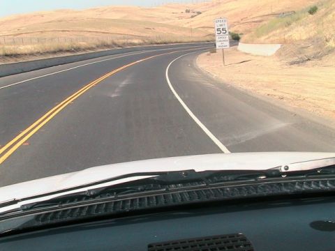 1971 Ford Bronco - View from inside a vehicle looking out at a highway curving through a dry, hilly landscape, with a speed limit sign visible.
