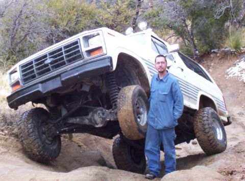 1983 Ford Bronco - A man poses next to a heavily modified white SUV, its off-road capabilities on a rocky trail.