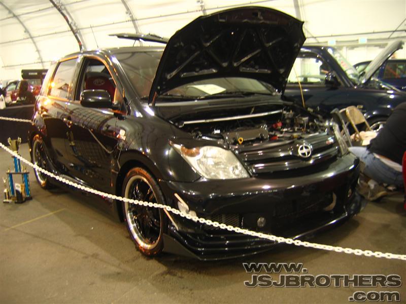 Black hatchback with hood open, showing engine bay. Another dark SUV is visible behind it