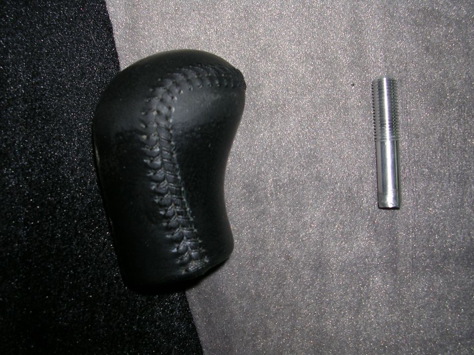 Black leather shift knob with visible stitching next to a threaded metal bolt on a textured gray surface