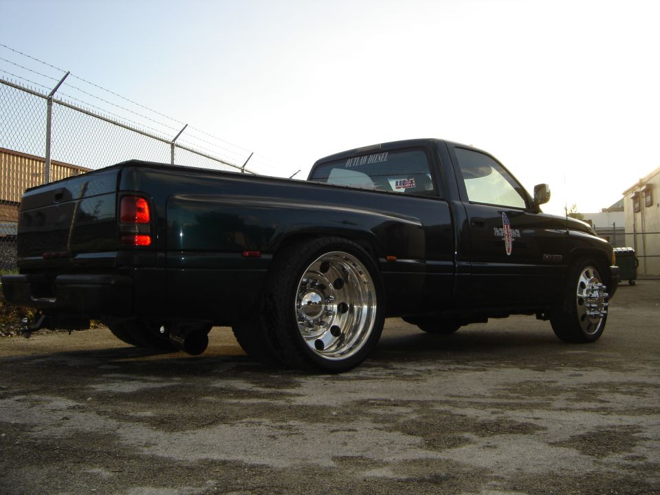 Dark green lowered Dodge Ram single cab pickup with polished dually-style wheels, rear 3/4 view by a barbed-wire fence