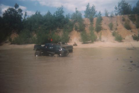2004 Honda TRX 450 FM - Two trucks navigate a muddy water crossing with a wooded area and sandy embankment.