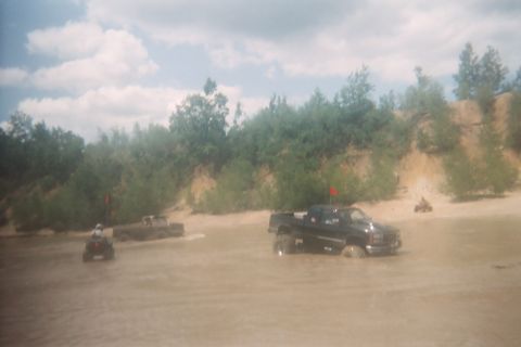 2004 Honda TRX 450 FM - Several vehicles are off-roading in a muddy, watery area with a wooded embankment under a cloudy sky.