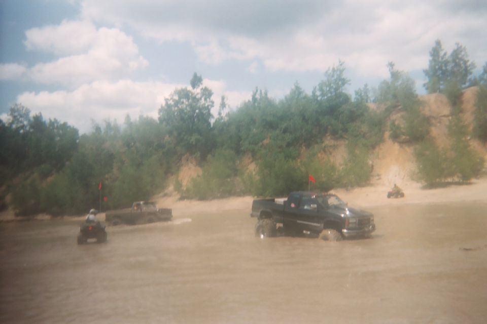 Black pickup truck and ATVs driving through muddy water near a sandy bank with trees