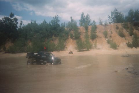 2004 Honda TRX 450 FM - A black pickup truck navigates through shallow, muddy water with a sandy embankment and trees in the background.