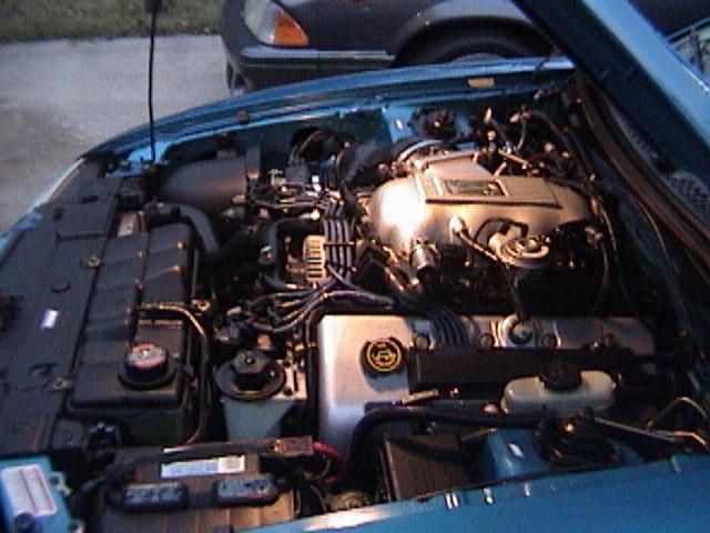Engine bay of a blue car, showing the motor and various hoses and components. Another car is partially visible in the background