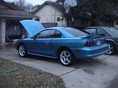 1989 Ford Mustang - A blue Ford Mustang is parked outside a garage with its hood up, next to another vehicle.