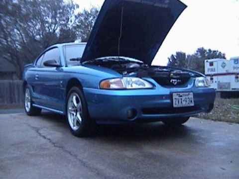 1989 Ford Mustang - The front end of a blue Ford Mustang is visible with its hood raised, revealing the engine bay.