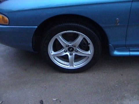 1989 Ford Mustang - A blue car is shown from the side, aftermarket wheels and a fender emblem.