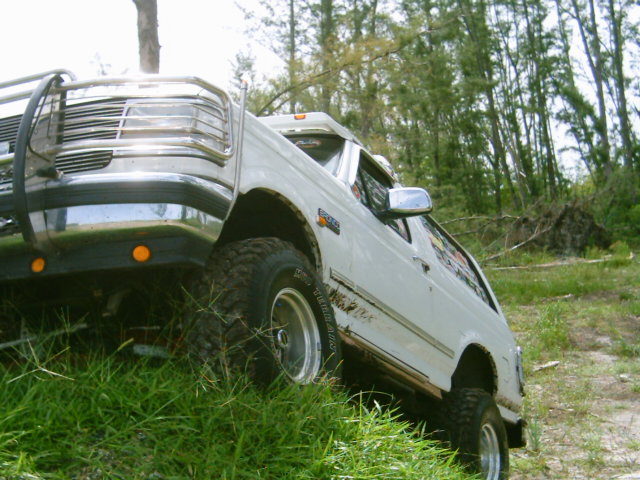 Low front-corner view of white lifted Ford Bronco with chrome brush guard on a grassy trail edge