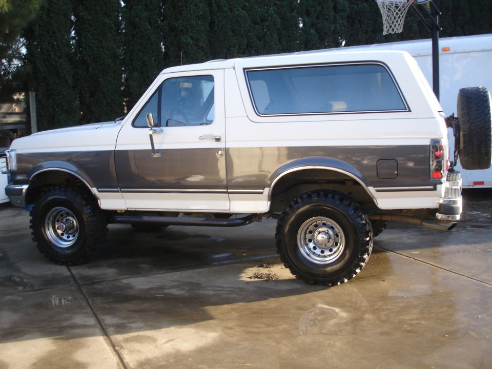 Lifted gray and white Ford Bronco on mud tires, driver-side profile on a concrete driveway