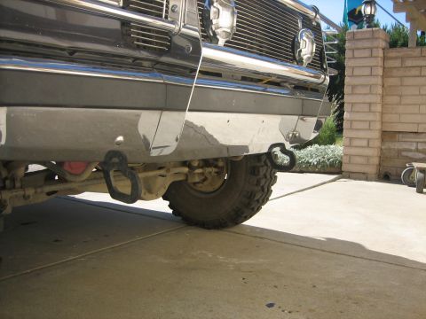1989 Ford Bronco - Low ground-level view under the front bumper of a lifted truck on a concrete driveway, showing a pair of black bolt-on tow hooks mounted to…