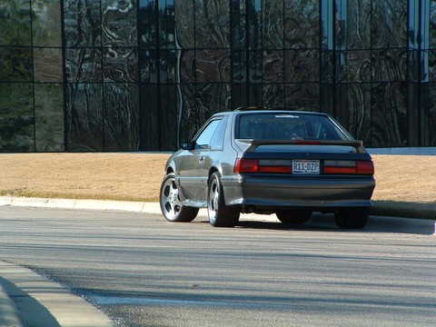 1989 Ford Mustang - A 1989 Ford Mustang is seen from the rear, parked in front of a modern building with a dark, reflective facade.