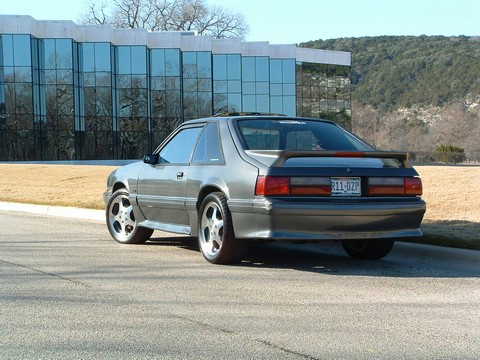 1989 Ford Mustang - A silver coupe with a rear spoiler is parked on a road in front of a modern building.