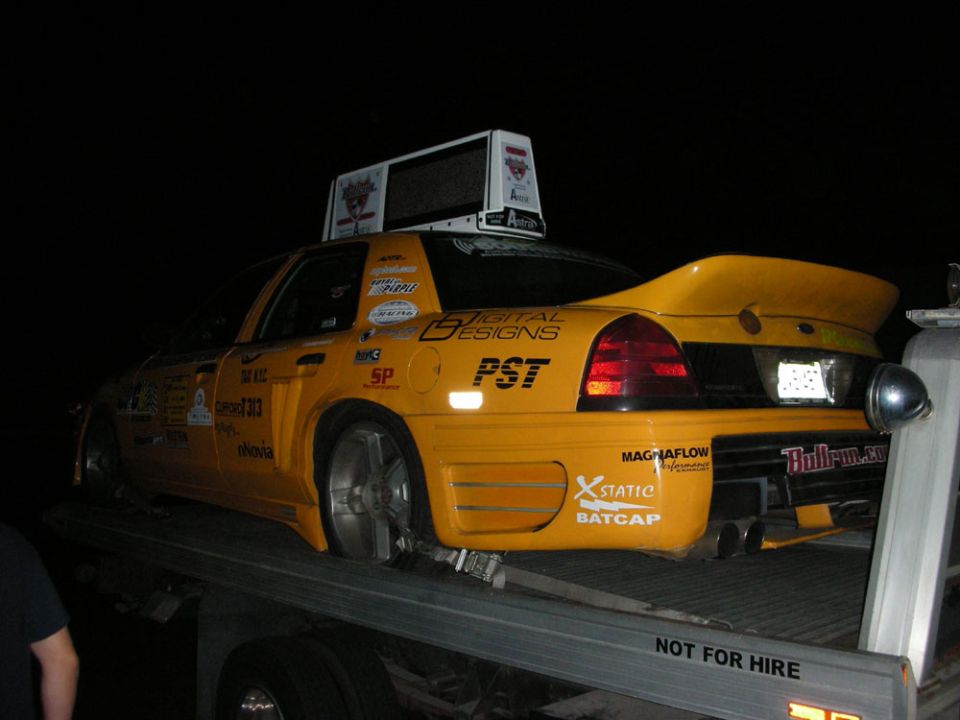 Yellow taxi with racing decals and a spoiler, on a flatbed truck at night. Bullrun.com visible